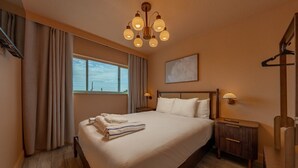 Ein Hotelzimmer mit einem großen Bett, einem Nachttisch, einer Lampe, einem Fenster mit Vorhängen und einer Deckenleuchte.