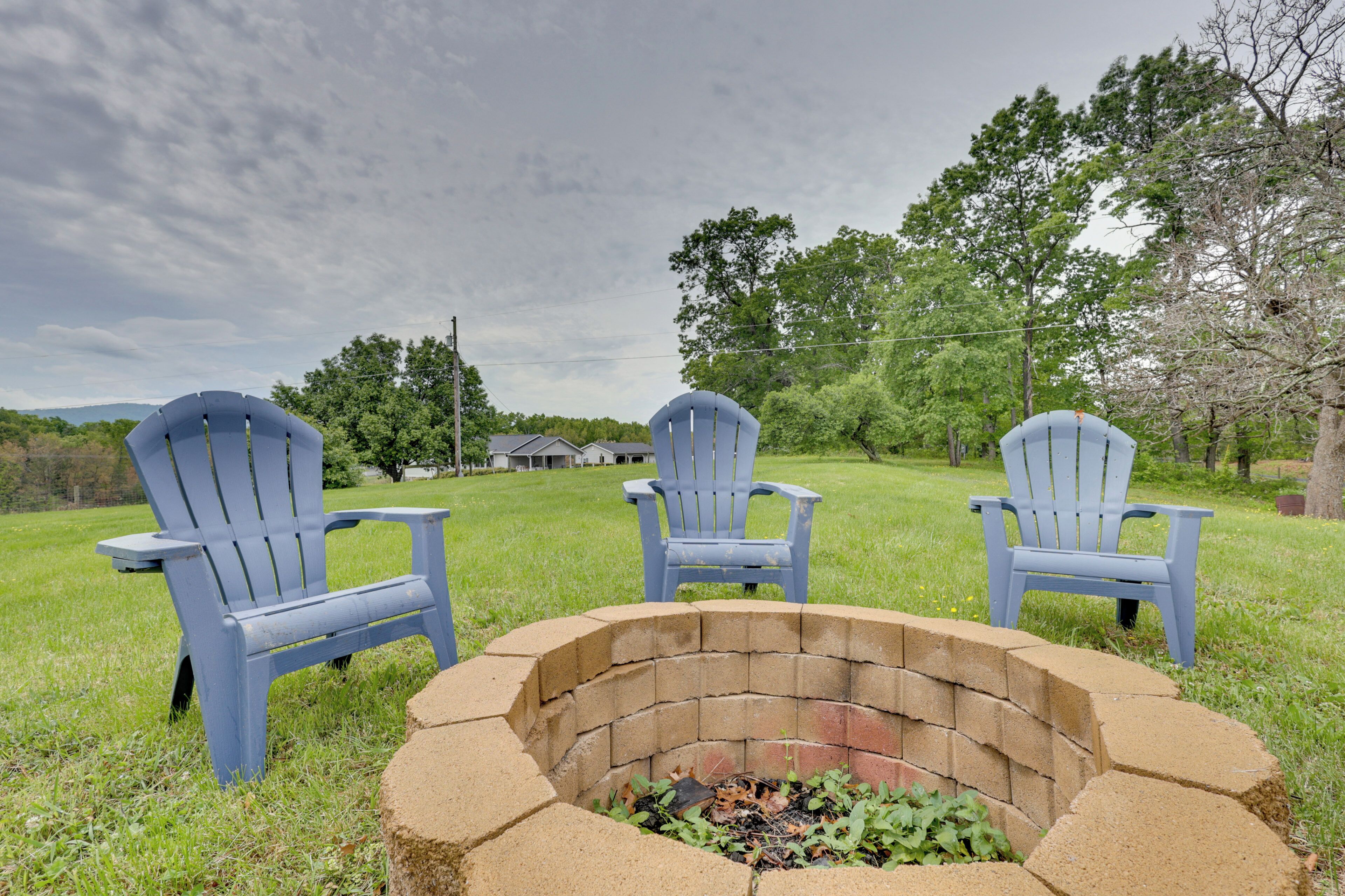 A grassy outdoor area with a brick fire pit and three blue Adirondack chairs.