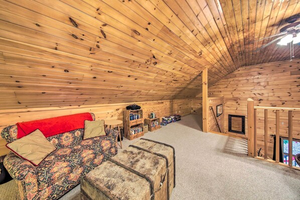 A cozy attic room with wooden walls and a sloped ceiling. It features a patterned sofa, a small table, and a suitcase.