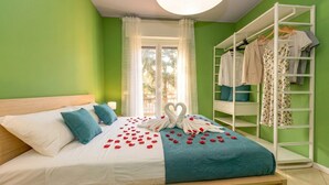 A neatly made bed with a heart-shaped towel arrangement and rose petals, a white wardrobe with hanging clothes, and a window with white curtains.