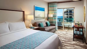 A hotel room with a large bed, a sofa, a desk, and a balcony with a view of the ocean.