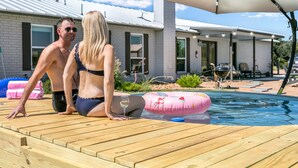 Una terraza de madera con piscina, un flotador rosa y una sombrilla blanca.