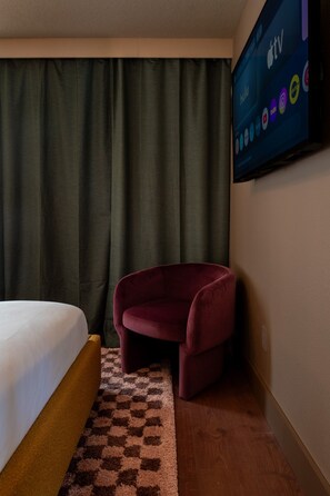 A hotel room with a bed, a red armchair, a checkered rug, and a TV displaying various channels.