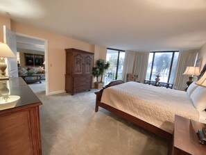 A bedroom with a large bed, a wooden dresser, a nightstand, a potted plant, and a view of the outdoors through the window.