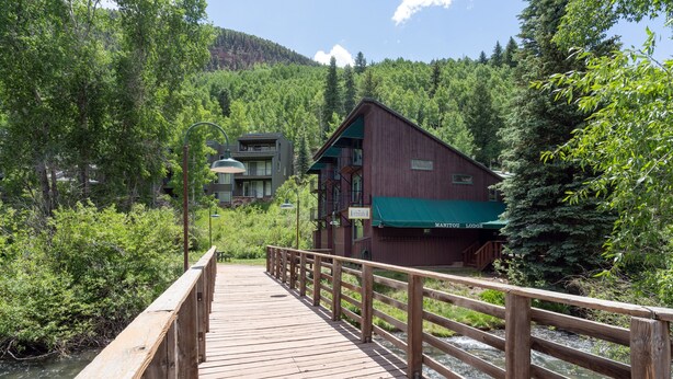 Uma ponte de madeira que leva a um chalé com telhado verde, cercado por uma floresta densa e um rio.
