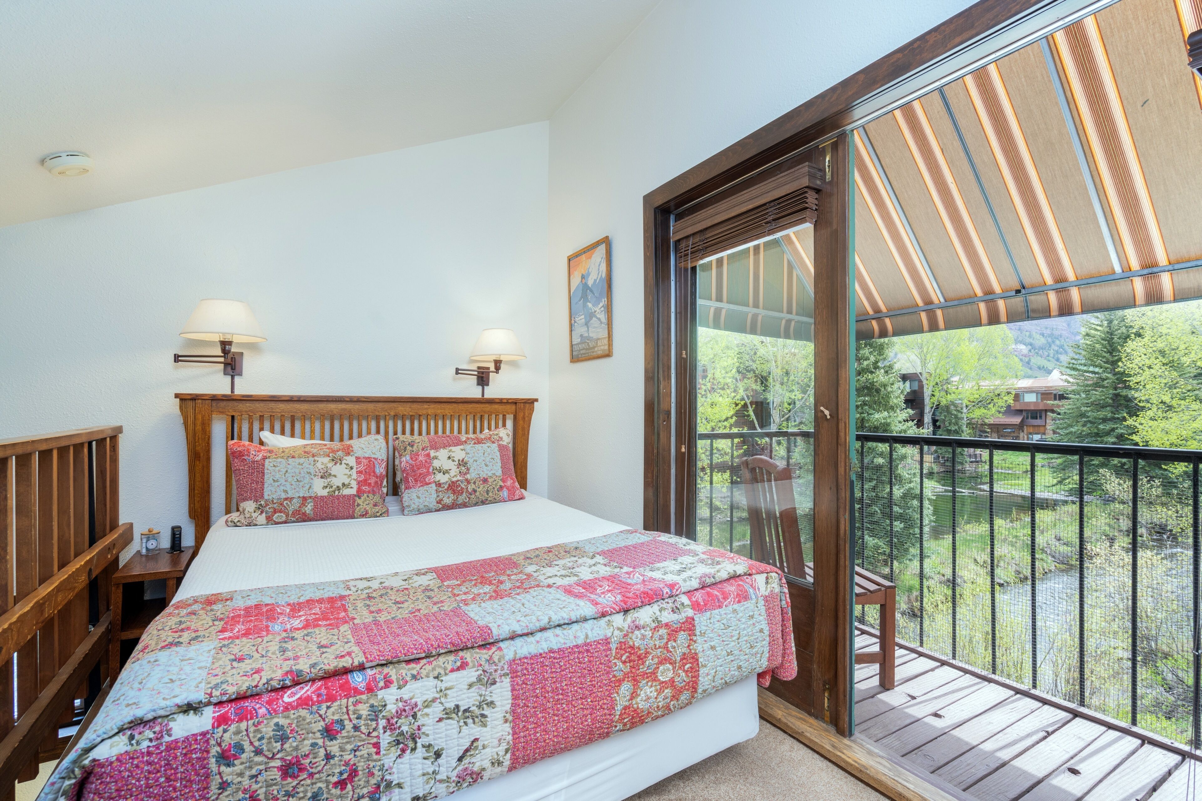 A bedroom with a large bed, two bedside lamps, a wooden headboard, a quilt, and a balcony with a view of greenery.