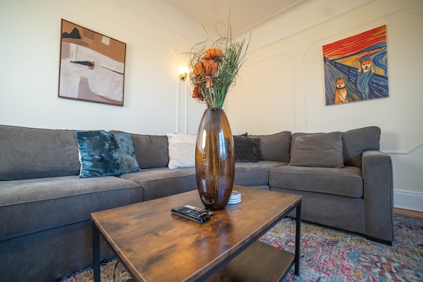 A living room with a sectional sofa, a coffee table, a vase with flowers, and two paintings on the walls.
