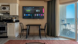 A modern living room with a flat-screen TV displaying Hulu and Apple TV apps, a kitchen with a microwave and stove, a dining area with a table and chairs, and a balcony with chairs. - Condo On The Beach - Ocean Side View Balcony (Belleair Beach)