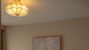 A neatly made bed with white linens, a headboard, and a patterned rug. There is a bedside table with a lamp, a framed artwork on the wall, and a ceiling light fixture. - Condo On The Beach - Ocean Side View Balcony (Belleair Beach)