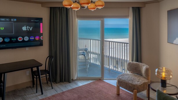 A hotel room with a TV displaying various streaming services, a balcony with a view of the beach, a dining table, and a cozy chair. - Condo On The Beach - Ocean Side View Balcony (Belleair Beach)