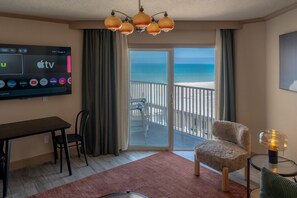 A hotel room with a TV displaying various streaming services, a balcony with a view of the beach, a dining table, and a cozy chair. - Condo On The Beach - Ocean Side View Balcony (Belleair Beach)