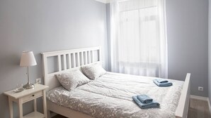 A bedroom with a bed, a nightstand, a lamp, and a window with sheer curtains. - Vintage Apartments Blue / Free parking (Rīga)
