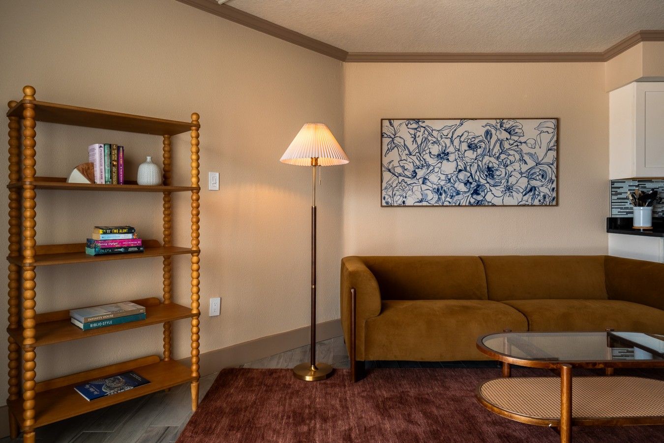 A living room with a wooden bookshelf, a brown sofa, a round coffee table, a floor lamp, and a floral artwork on the wall.