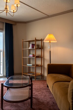 Une chambre d’hôtel avec une table à plateau en verre, un canapé marron, une bibliothèque en bois et un lampadaire.