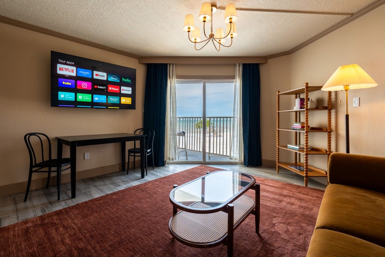 A hotel room with a TV displaying various streaming apps, a dining table with chairs, a glass coffee table, a sofa, a bookshelf, and a balcony with a view.