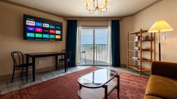 A hotel room with a TV displaying various streaming apps, a dining table with chairs, a glass coffee table, a sofa, a bookshelf, and a balcony with a view.