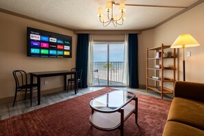A hotel room with a TV displaying various streaming apps, a dining table with chairs, a glass coffee table, a sofa, a bookshelf, and a balcony with a view. - Dreamy Sunset Condo - Side Ocean View Balcony (Belleair Beach)