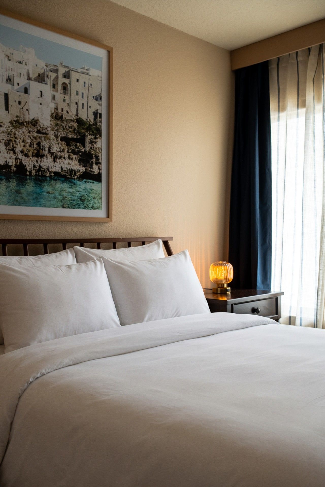 A hotel room with a large bed, a nightstand with a lamp, a framed picture on the wall, and a window with curtains.