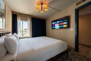 A hotel room with a large bed, a flat-screen TV displaying various streaming services, a window with curtains, and a chandelier. - Dreamy Sunset Condo - Side Ocean View Balcony (Belleair Beach)