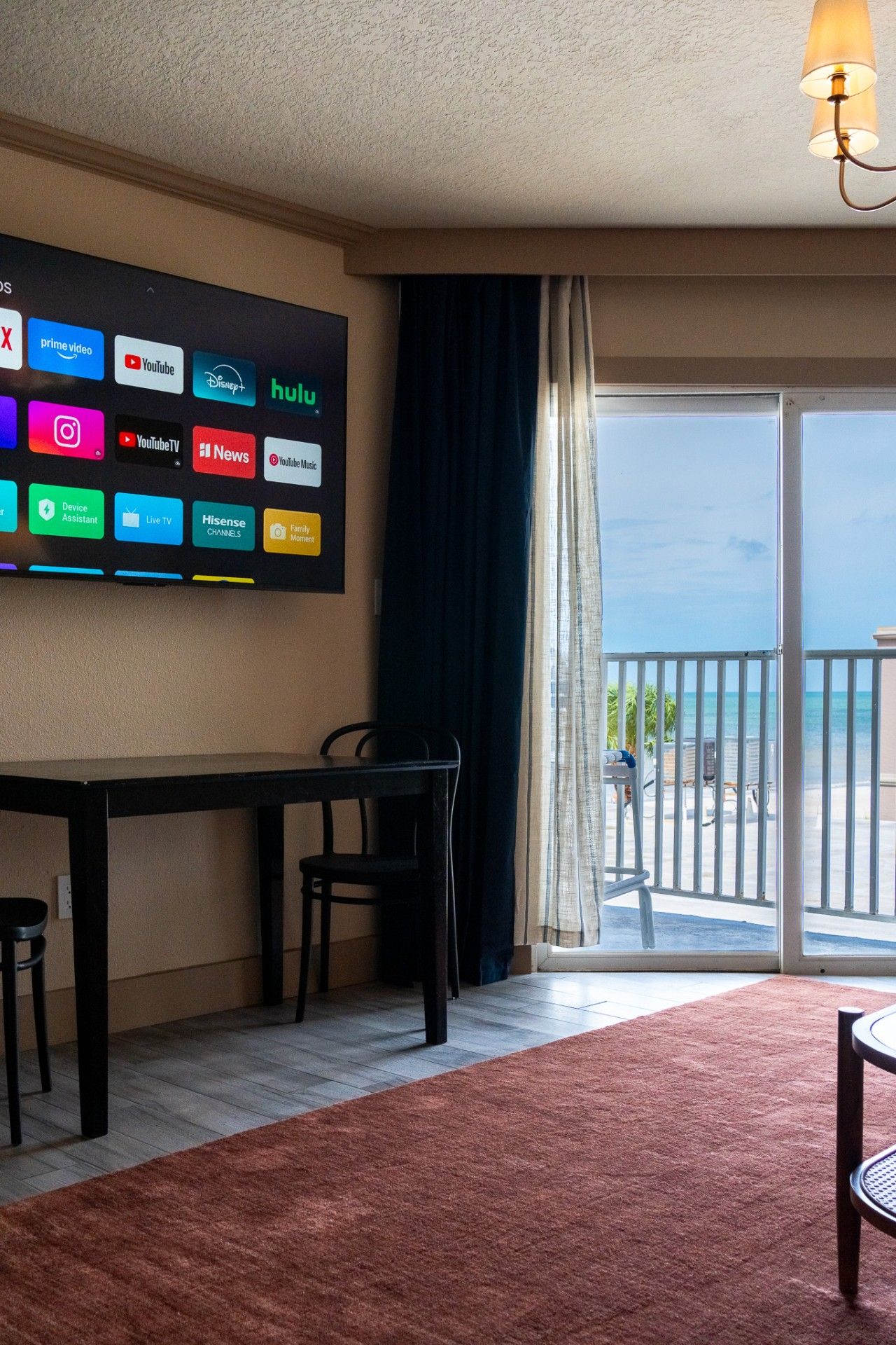 A hotel room with a flat-screen TV displaying various streaming services, a dining table with chairs, a red carpet, and a balcony with a view of the ocean.