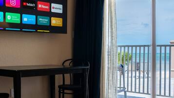 A hotel room with a flat-screen TV displaying various streaming services, a dining table with chairs, a red carpet, and a balcony with a view of the ocean.