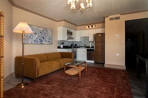 A living room with a sofa, a coffee table, and a lamp. There is a kitchen in the background with a refrigerator and microwave.