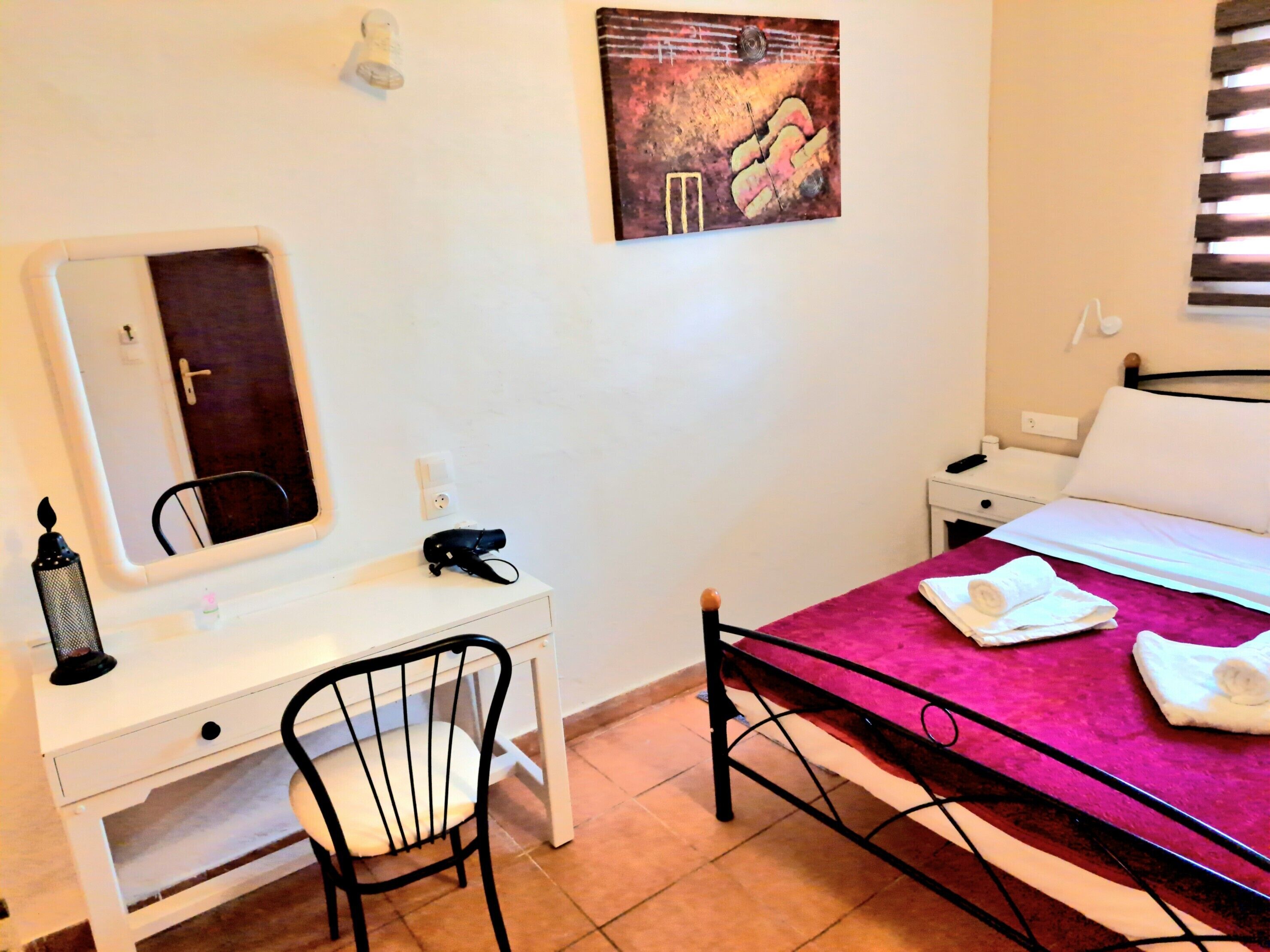A hotel room with a bed, a desk with a chair, a mirror, a nightstand, and a wall-mounted artwork.