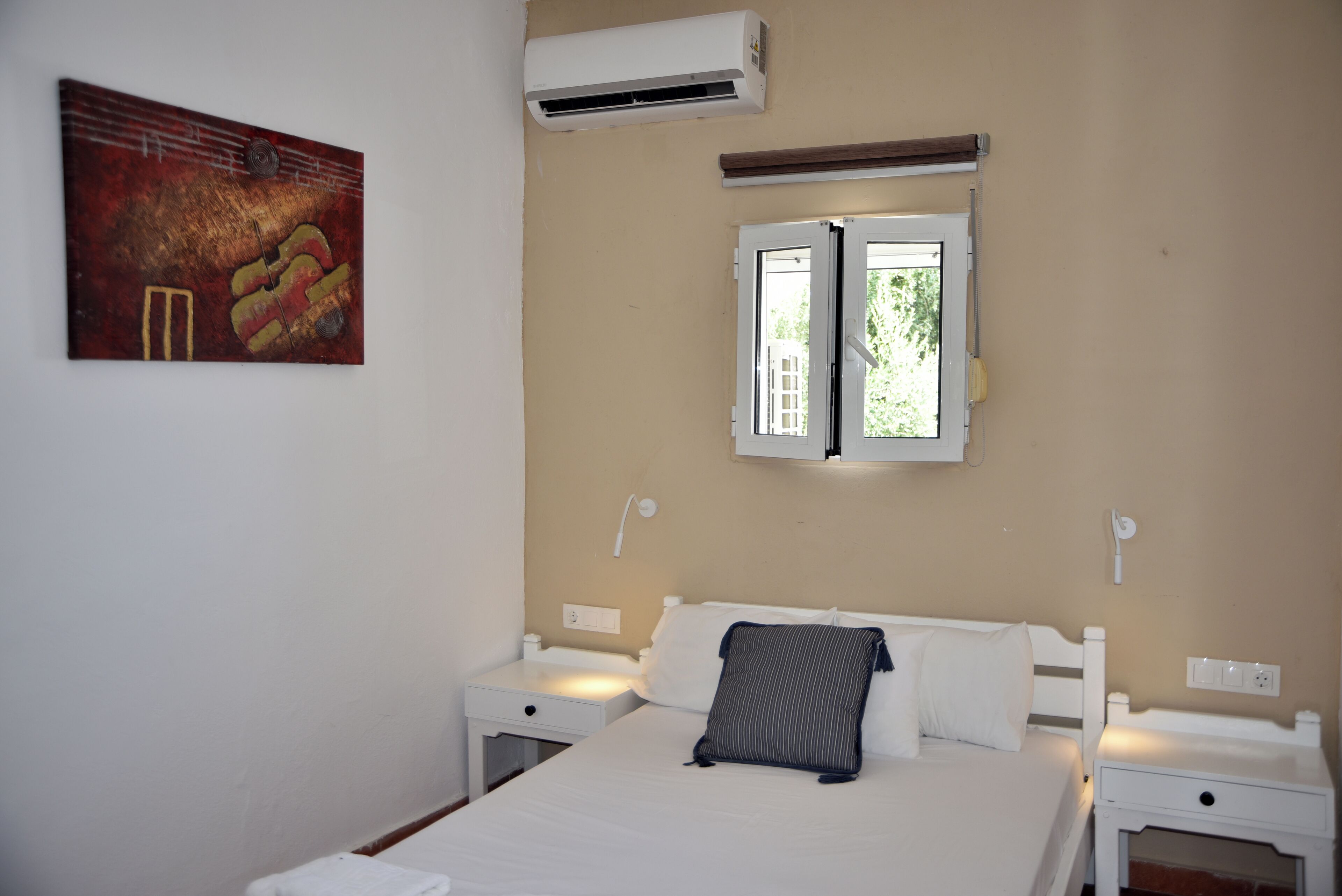 A bedroom with a bed, two bedside tables, an air conditioner, a window, and a painting on the wall.
