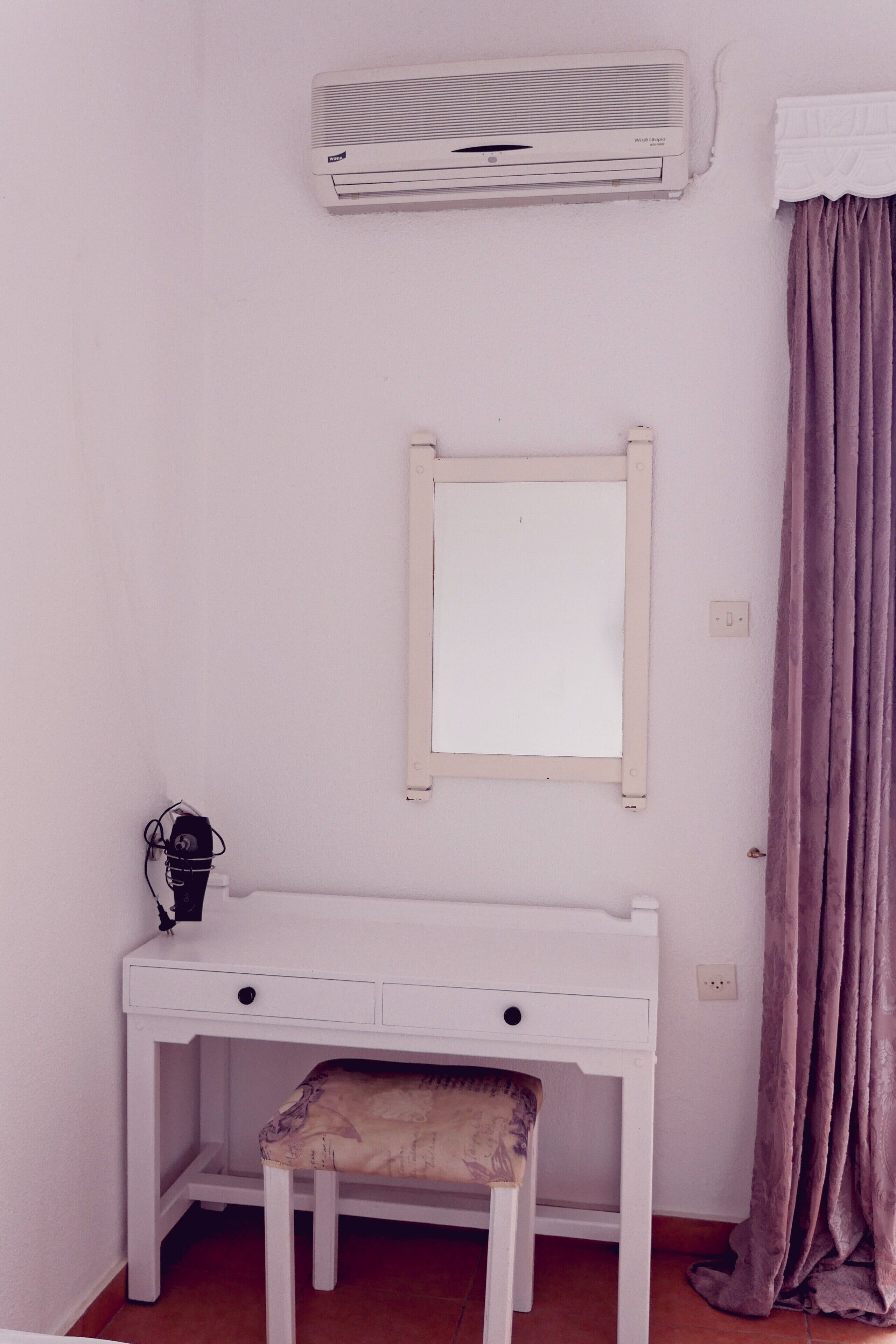 Un escritorio blanco con un espejo, un taburete pequeño y un aire acondicionado en la pared.