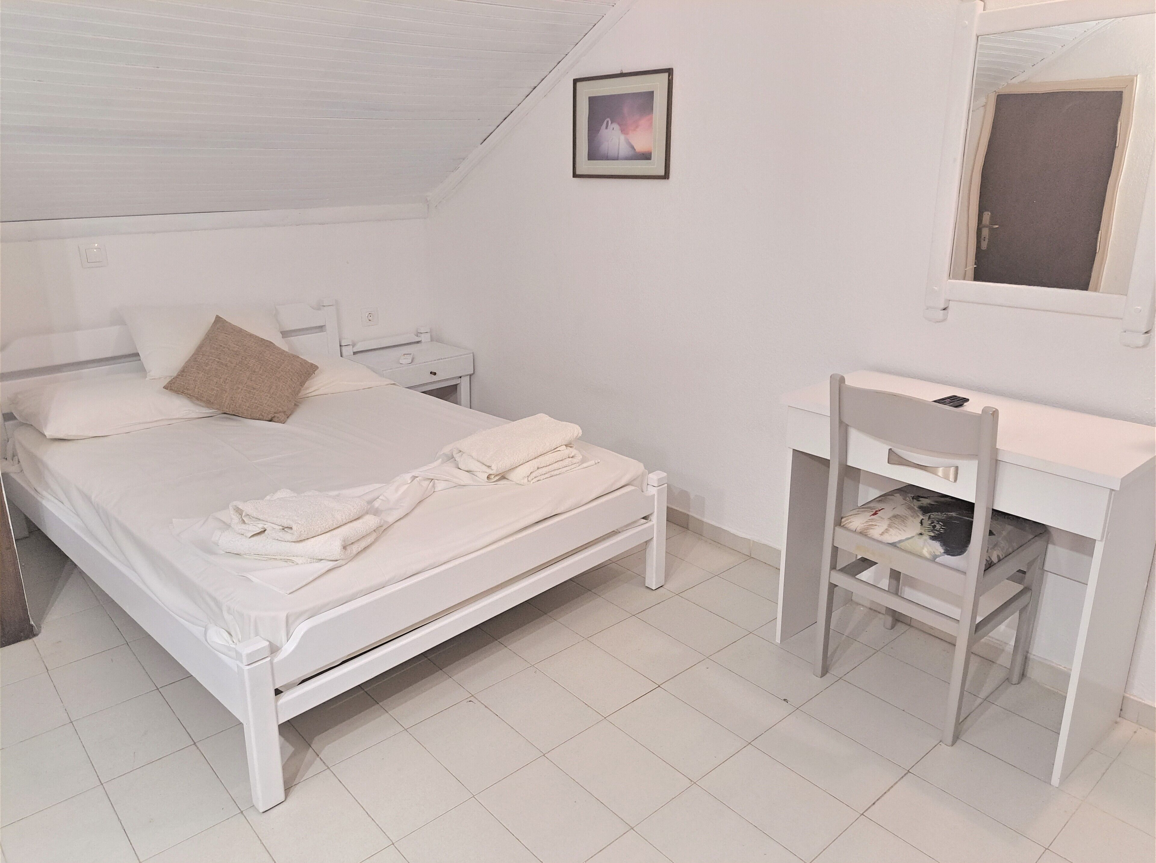 Een witte slaapkamer met een bed, nachtkastje, stoel en bureau met stoel.