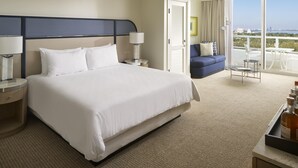 Quarto de hotel com uma cama grande, duas mesas de cabeceira com luminárias, um sofá, uma varanda com vista e uma pequena mesa com garrafas.