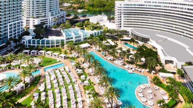 Um grande hotel com uma piscina central cercada por espreguiçadeiras e palmeiras.
