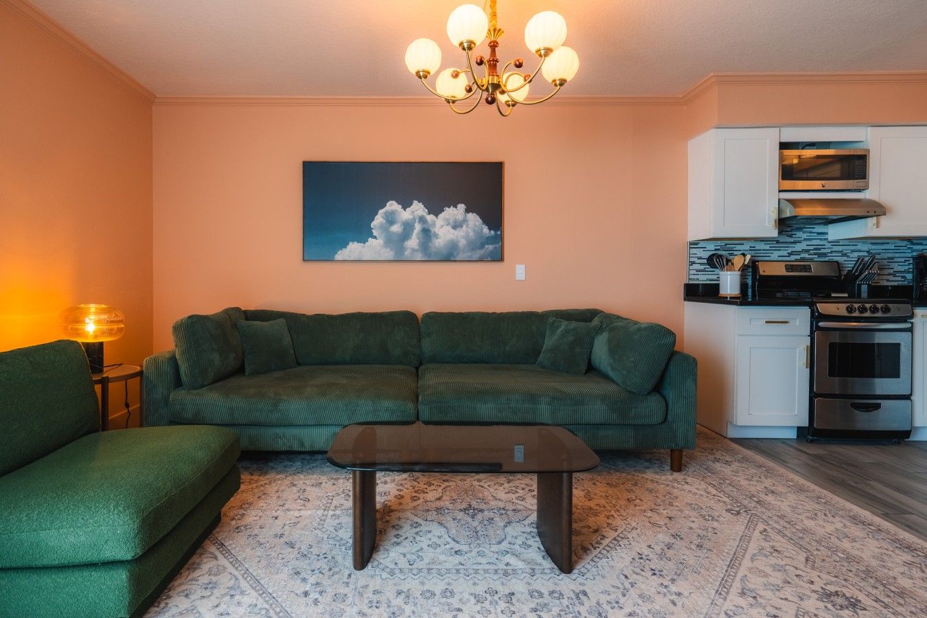 A living room with a green sofa, a coffee table, a chandelier, a painting of clouds, and a kitchen with modern appliances.