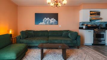 A living room with a green sofa, a coffee table, a chandelier, a painting of clouds, and a kitchen with modern appliances.