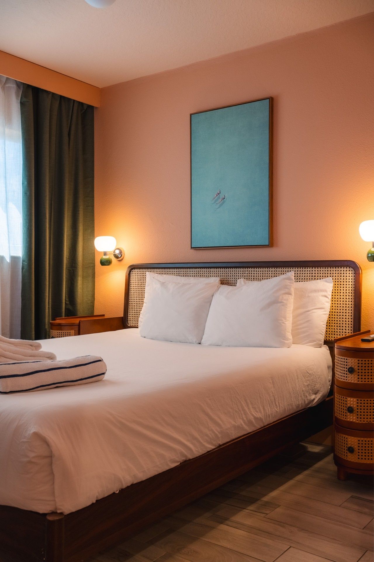 A neatly made bed with white linens and pillows, a single bedside table with a lamp, and a painting above the bed.