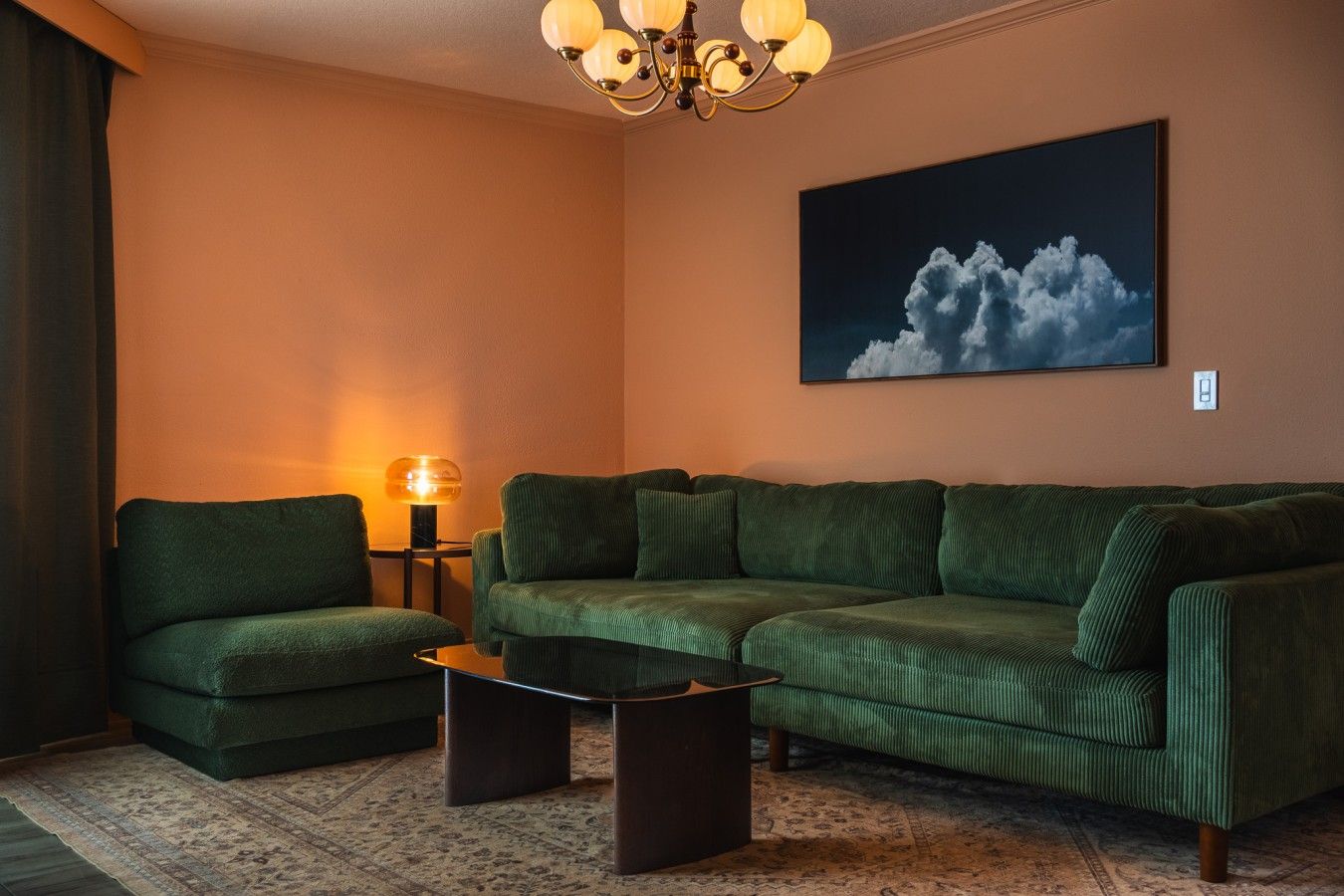 A living room with a sectional sofa, a coffee table, a lamp, and a painting of clouds on the wall.