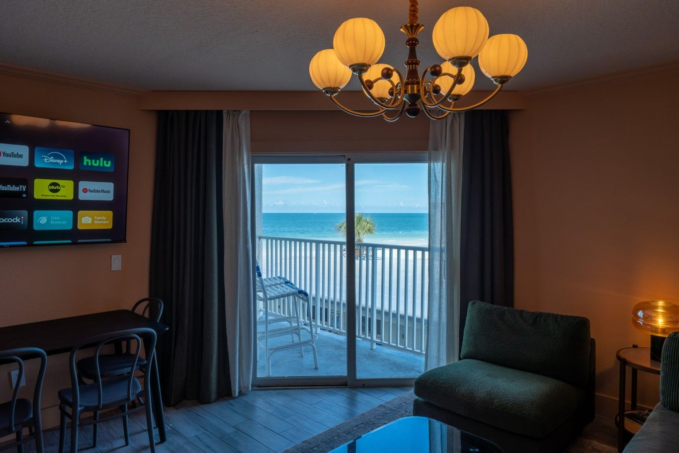 Ein Hotelzimmer mit Balkon, einem Esstisch mit Stühlen, einer Couch und einem Fernseher, der verschiedene Streaming-Dienste anzeigt.