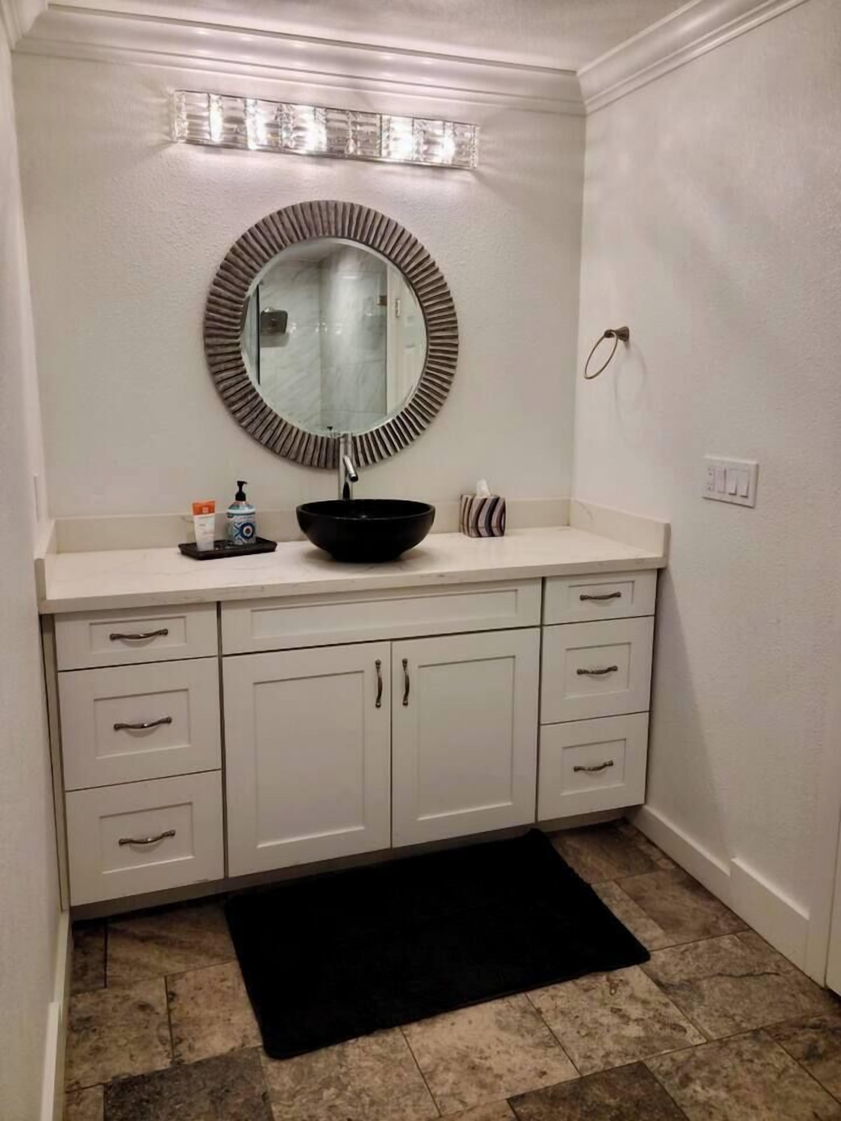 A bathroom with a round mirror, a black sink, and white cabinets.
