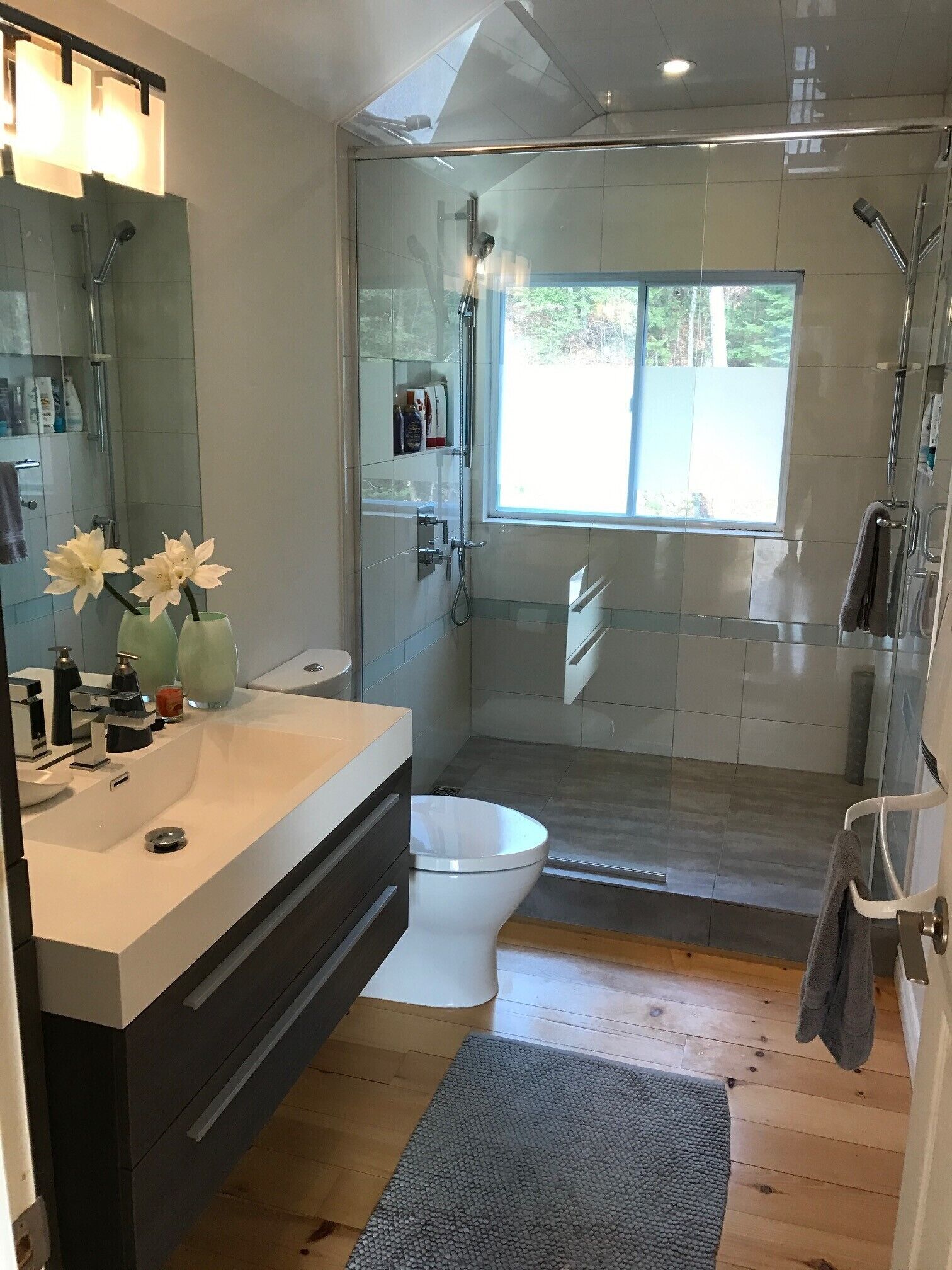 Une salle de bain moderne avec une douche à l’italienne en verre, des toilettes blanches et un meuble-lavabo agrémenté d’un miroir et de deux vases de fleurs.
