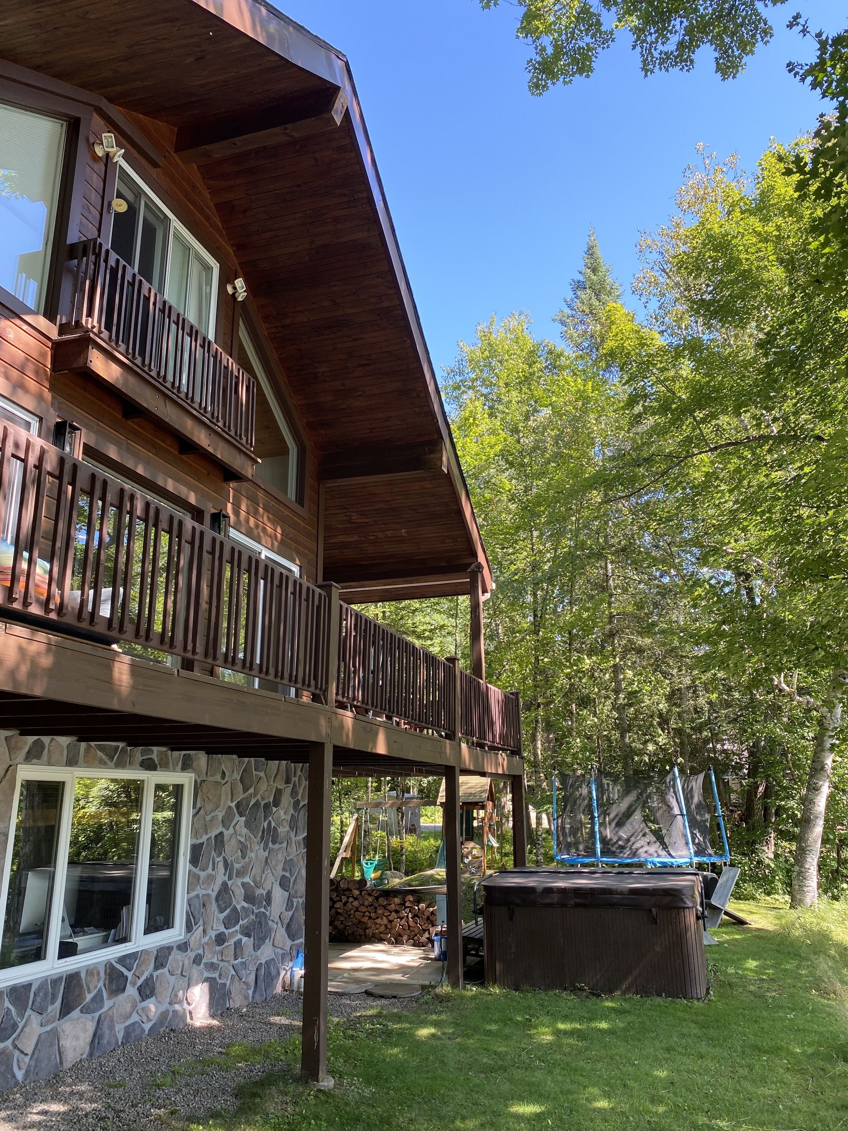 Une maison en bois à deux étages, avec des fondations en pierre, un balcon et une cour arrière dotée d’un trampoline.