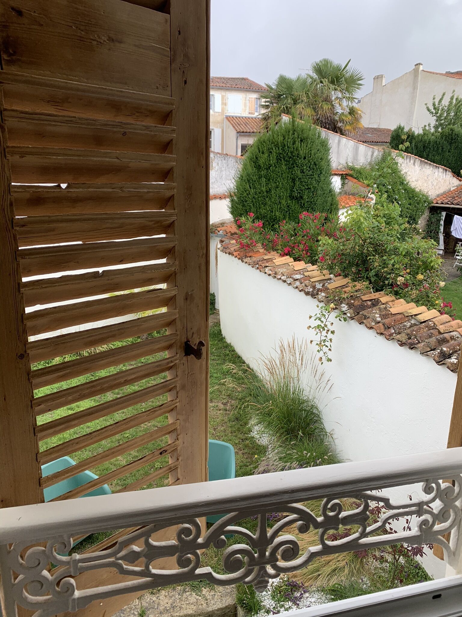 A wooden balcony with a white railing, overlooking a garden with various plants and a white wall.