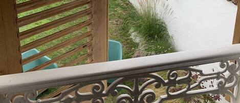 A wooden balcony with a white railing, overlooking a garden with various plants and a white wall.