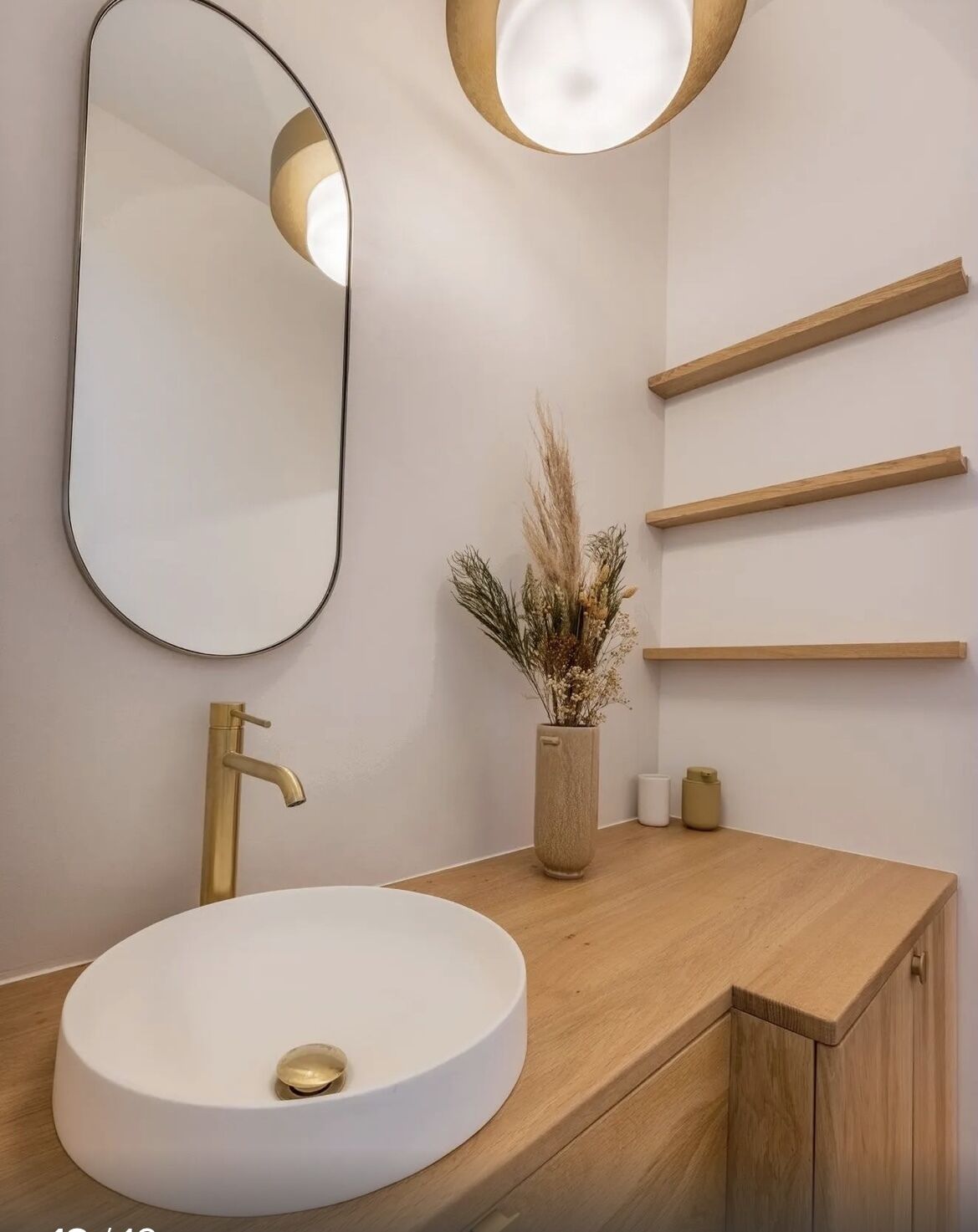 A modern bathroom with a round white sink, a gold faucet, a large mirror, and wooden shelves.