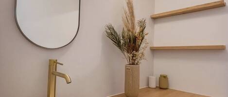 A modern bathroom with a round white sink, a gold faucet, a large mirror, and wooden shelves.