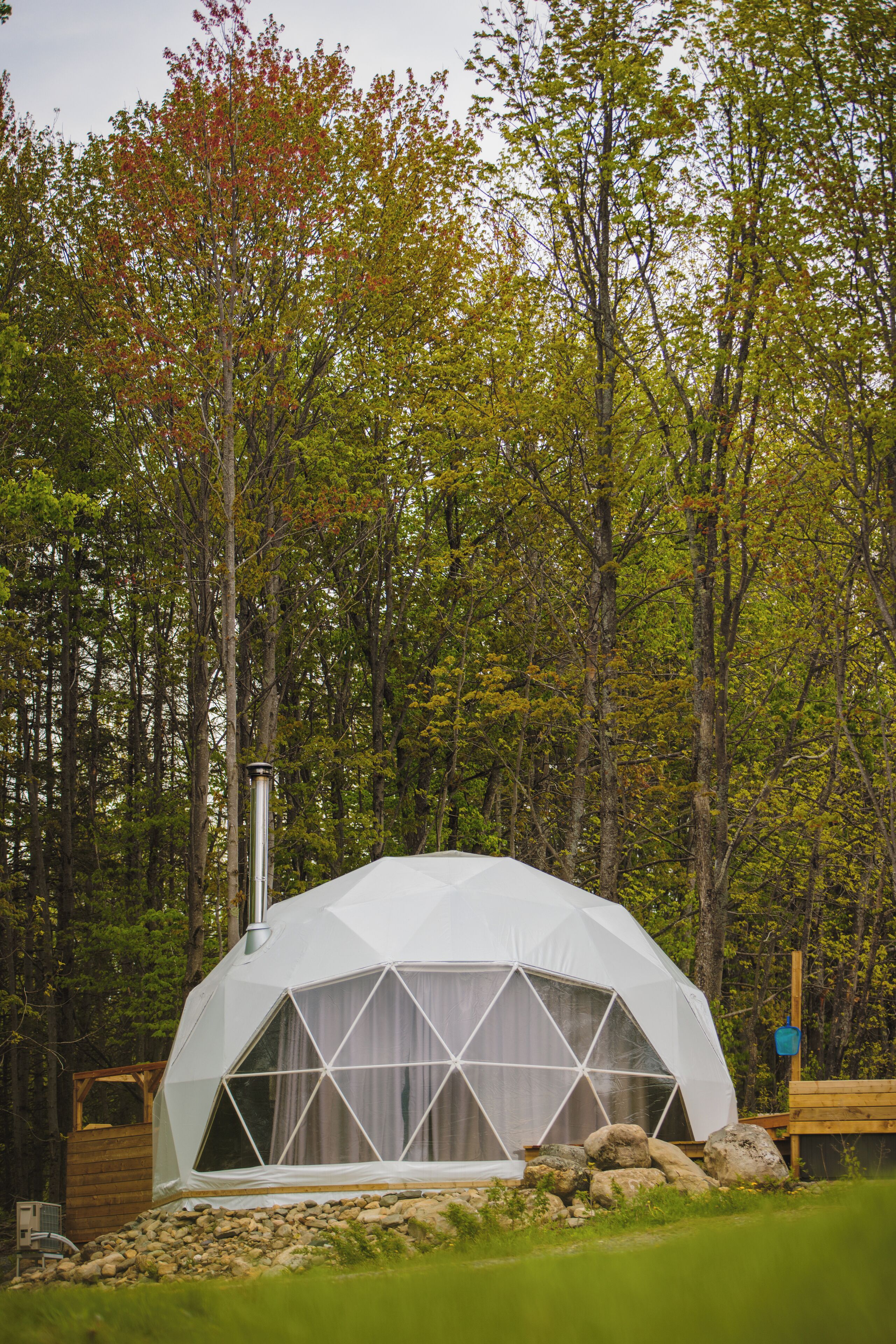 A geodesic dome structure in a forest setting with a chimney, surrounded by rocks and trees.