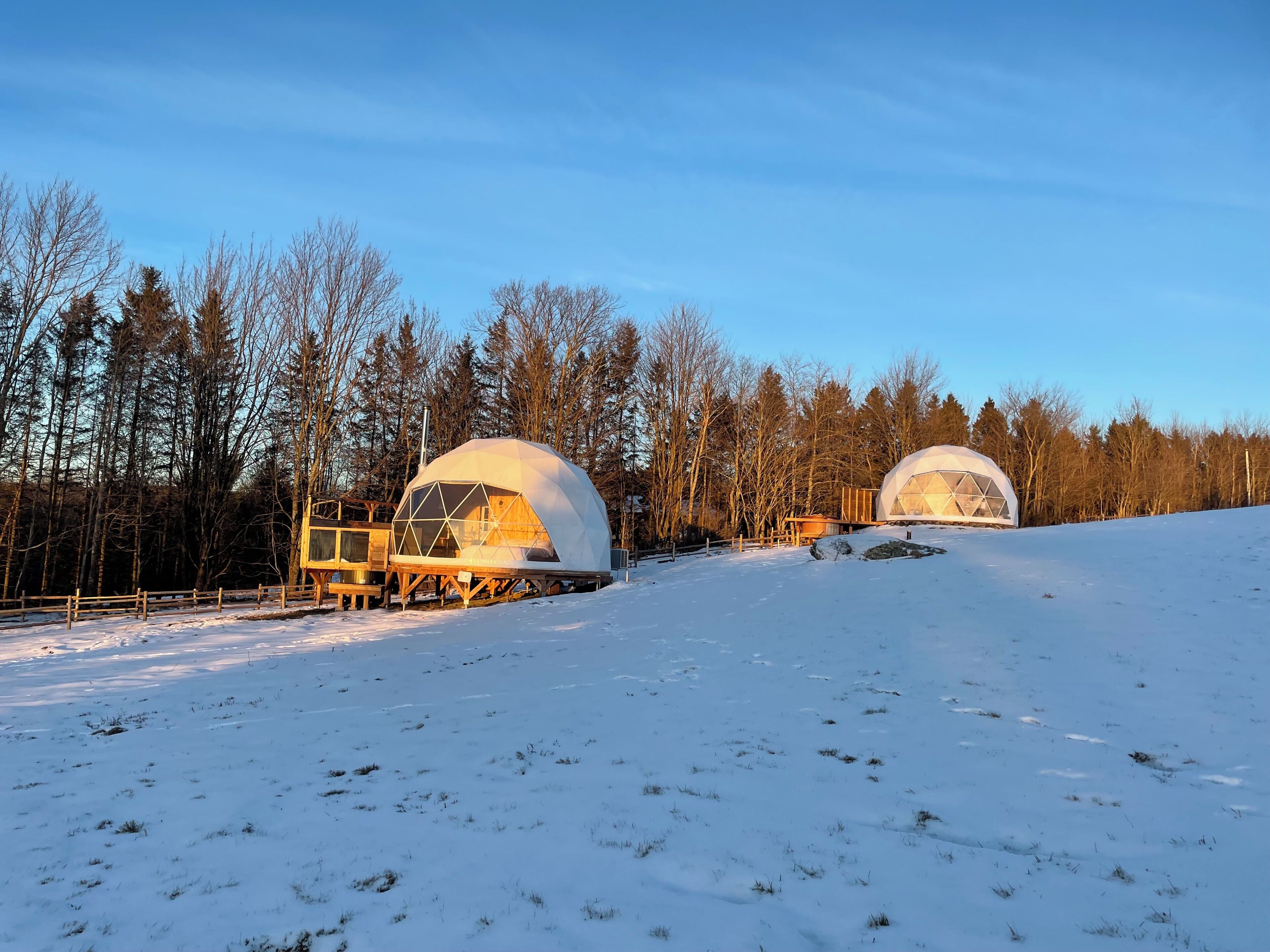 Two geodesic dome cabins in a snowy landscape with a wooden deck and a small building nearby.
