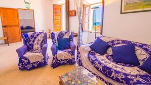Une chambre avec un lit, deux fauteuils à motifs, une petite table et une vue sur la mer par une porte ouverte.