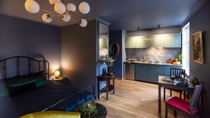 Une chambre à coucher de taille réduite, comprenant un lit, une table de chevet et un lustre moderne. À côté se trouve une petite cuisine équipée avec des placards et un coin repas avec une table et des chaises.