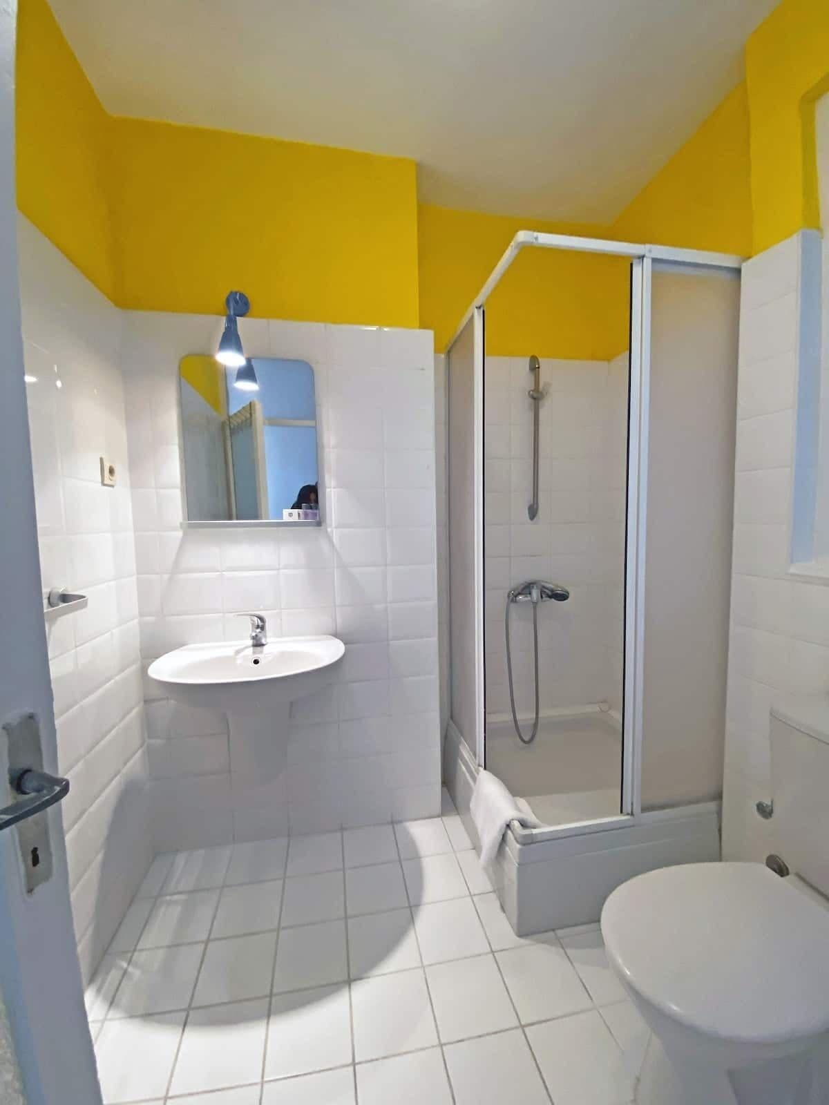 Une salle de bain avec un lavabo blanc, un miroir et des toilettes, une douche en verre et des murs jaunes.