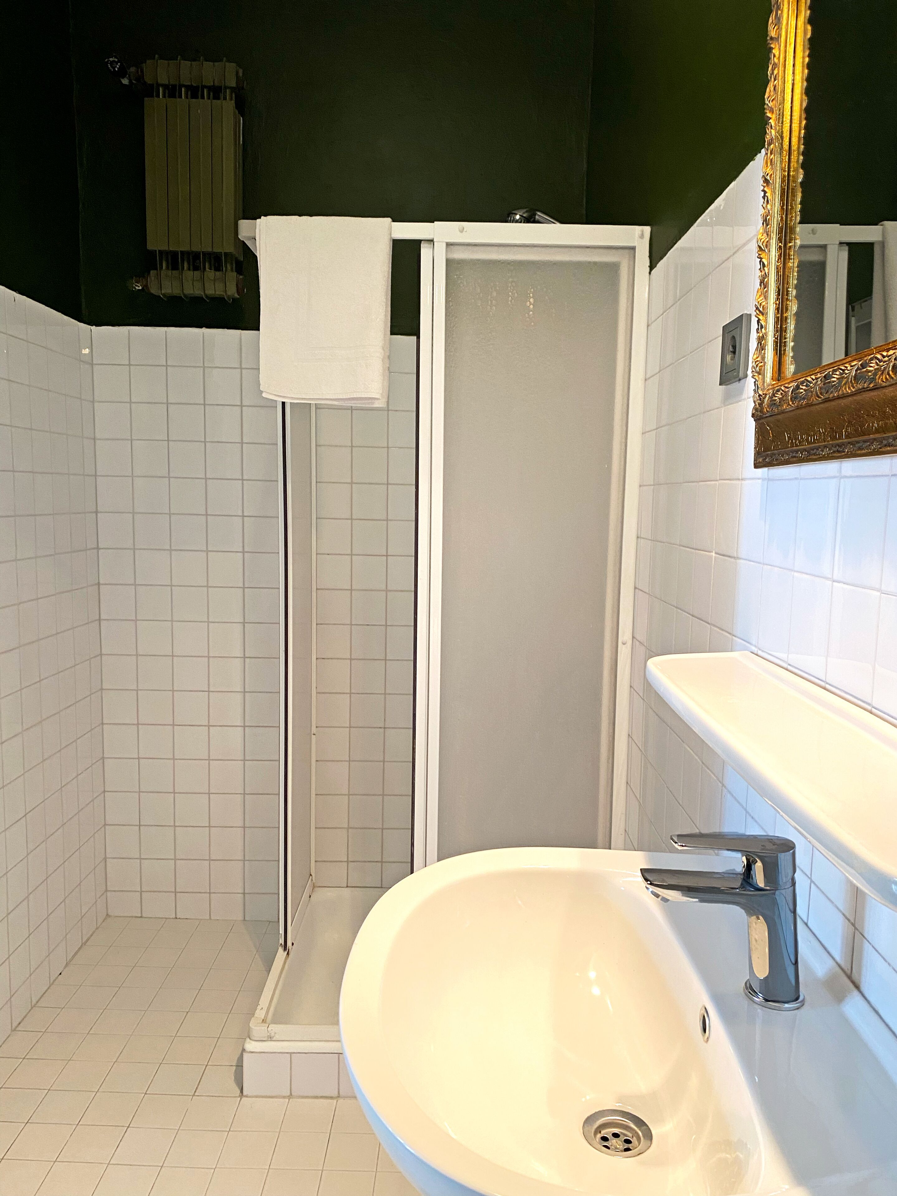 Une salle de bain avec un lavabo blanc, un miroir doté d’un cadre orné et un espace douche séparé par une porte en verre.